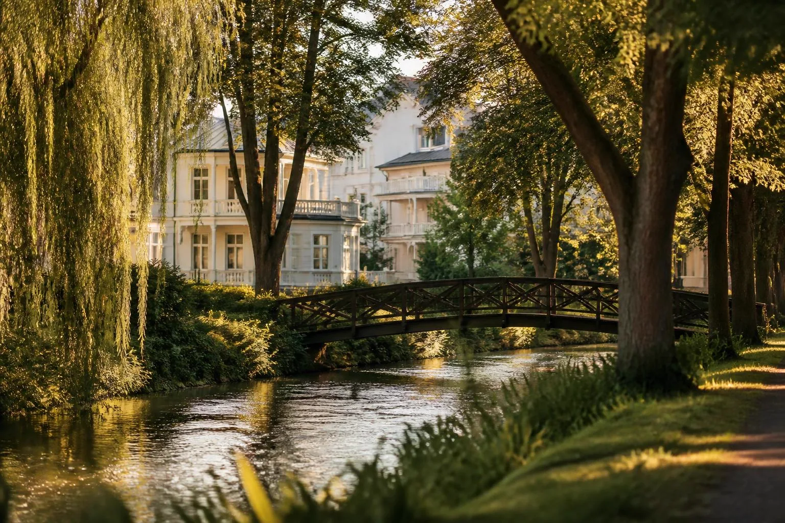Kleiner Fluss mit Trauerweiden und hölzerner Bogenbrücke, weiße Villen zwischen alten Bäumen