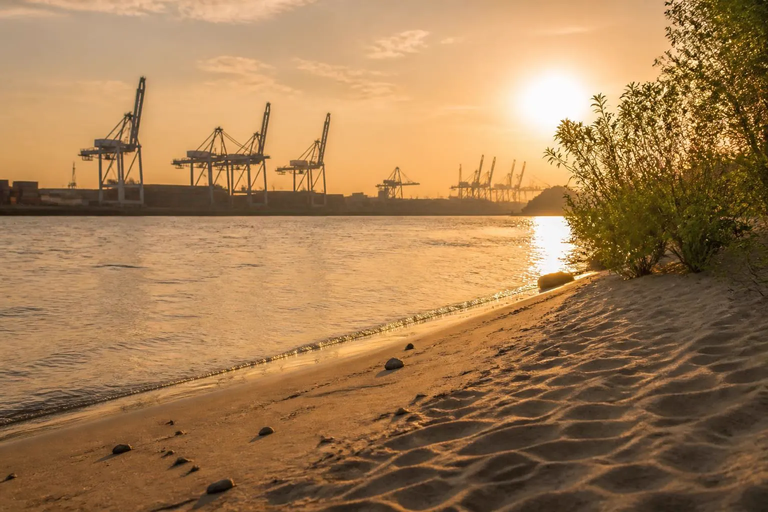Sandiger Elbstrand im Gegenlicht, am anderen Ufer Silhouetten von Hafenkränen