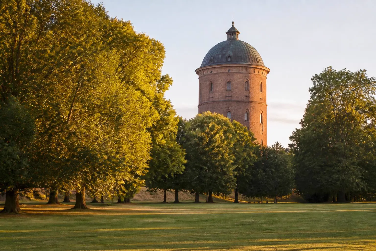 Weite Parkwiese mit alten Bäumen, dahinter ein runder Backsteinturm mit Kuppel