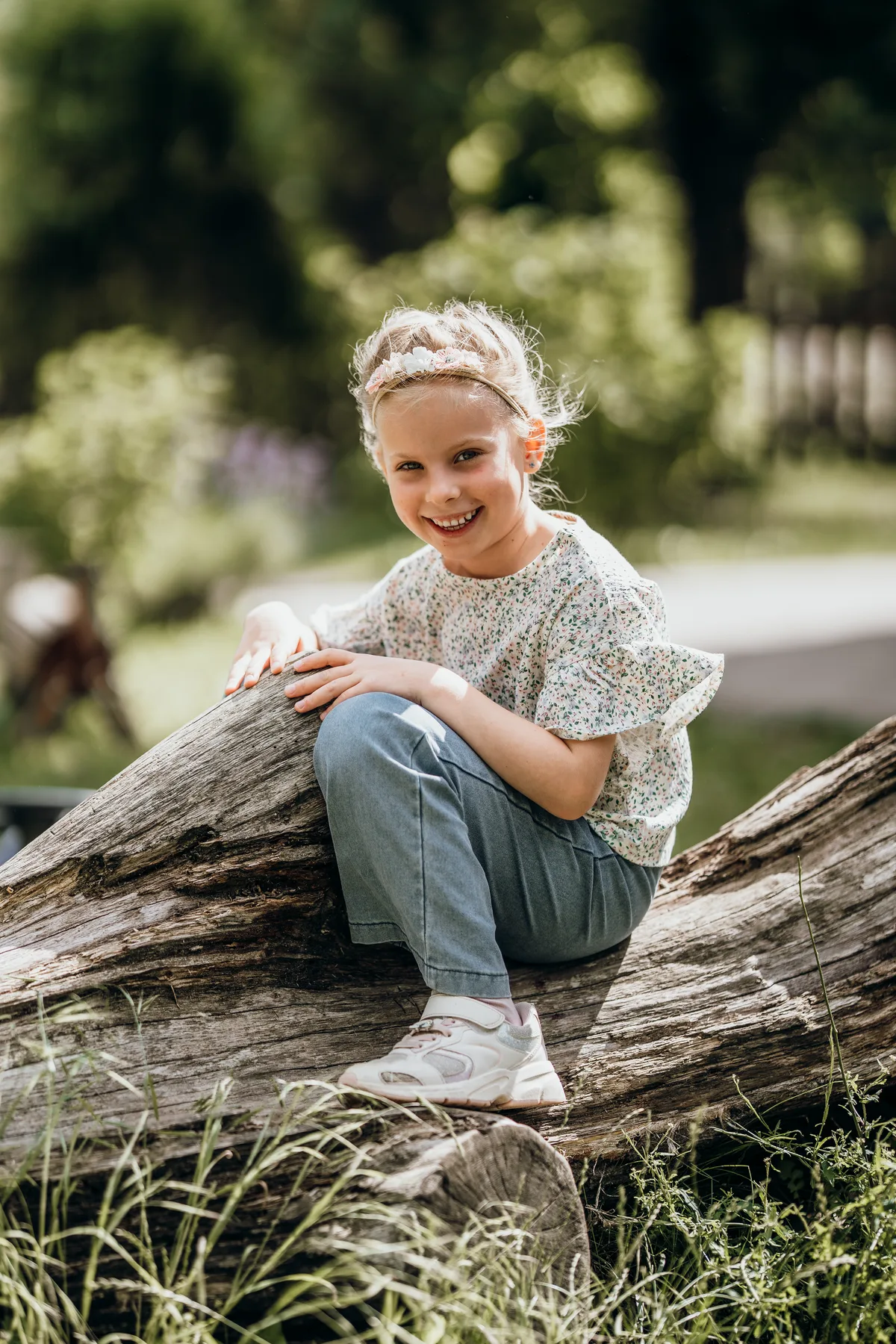 Kindergartenfotografie: Mädchen sitzt lachend auf einem liegenden Baumstamm im Grünen