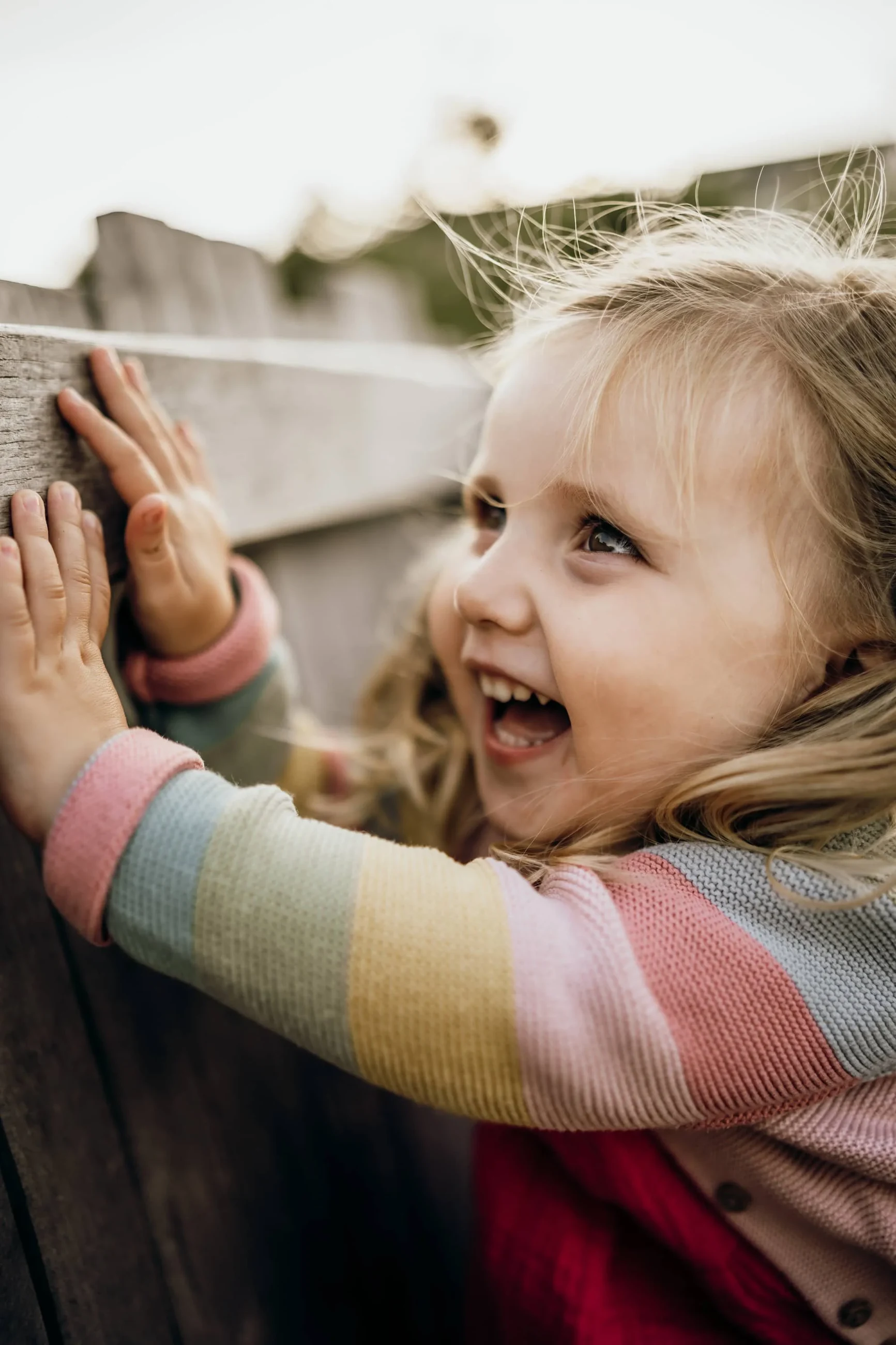 Kindergartenfotografie: Kleinkind im Regenbogen-Pullover lacht am Holzzaun im Kita-Garten