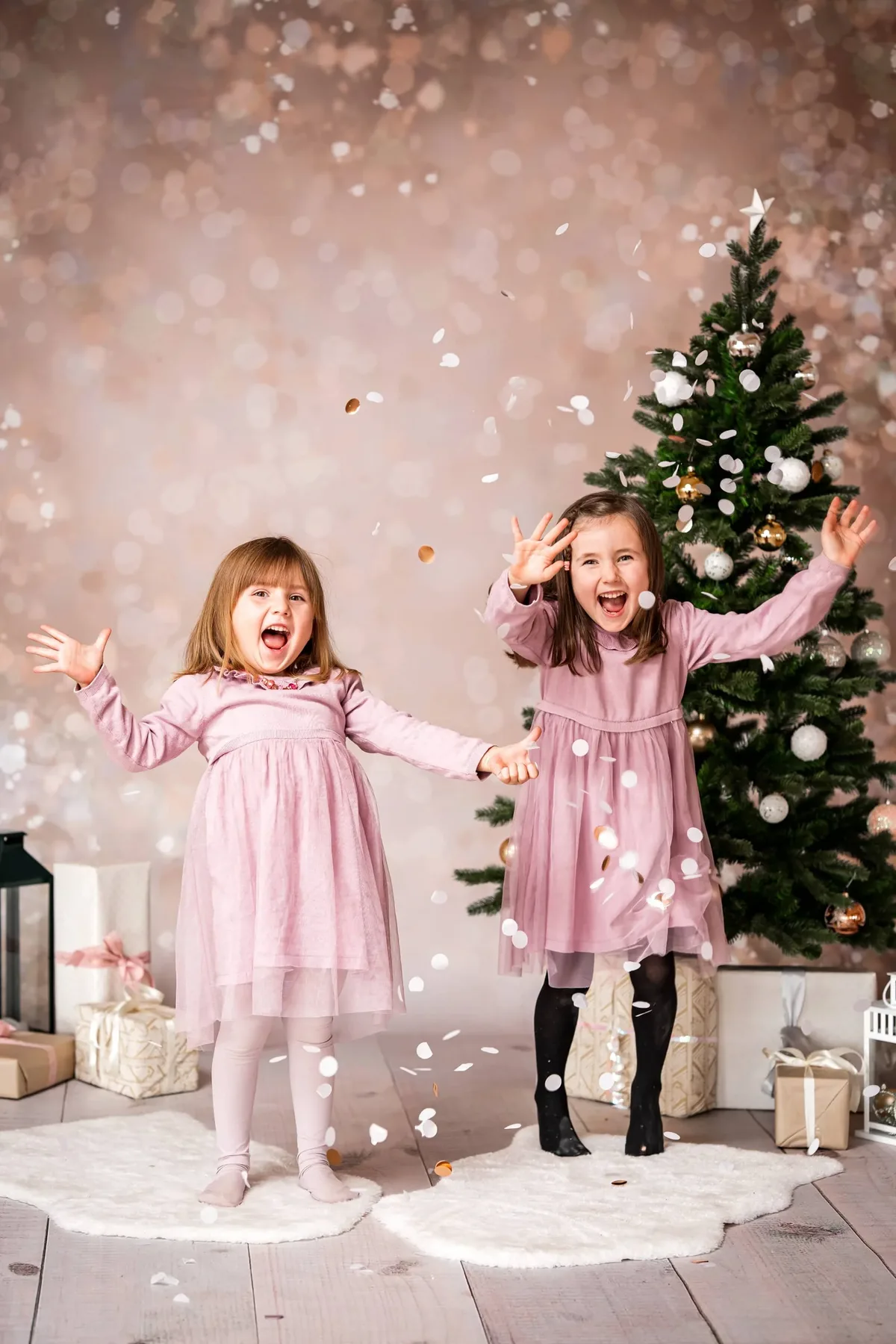 Kinderfotografie: Zwei Mädchen in rosa Kleidern jubeln im Konfettiregen vor dem Weihnachtsbaum