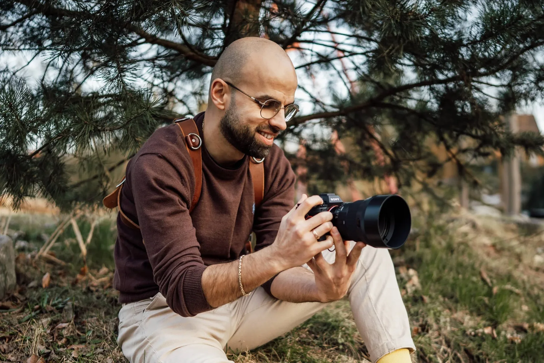 Fotograf hockt mit Kamera unter einer Kiefer und lächelt in die Kamera