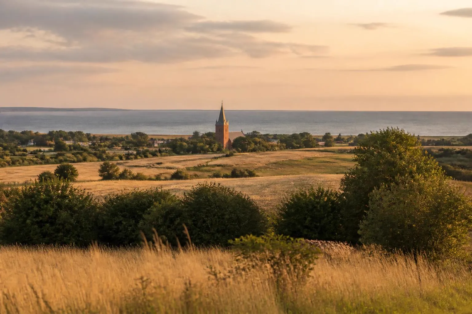 Blick über Felder zur Ostsee, dazwischen der Turm einer kleinen Backsteinkirche
