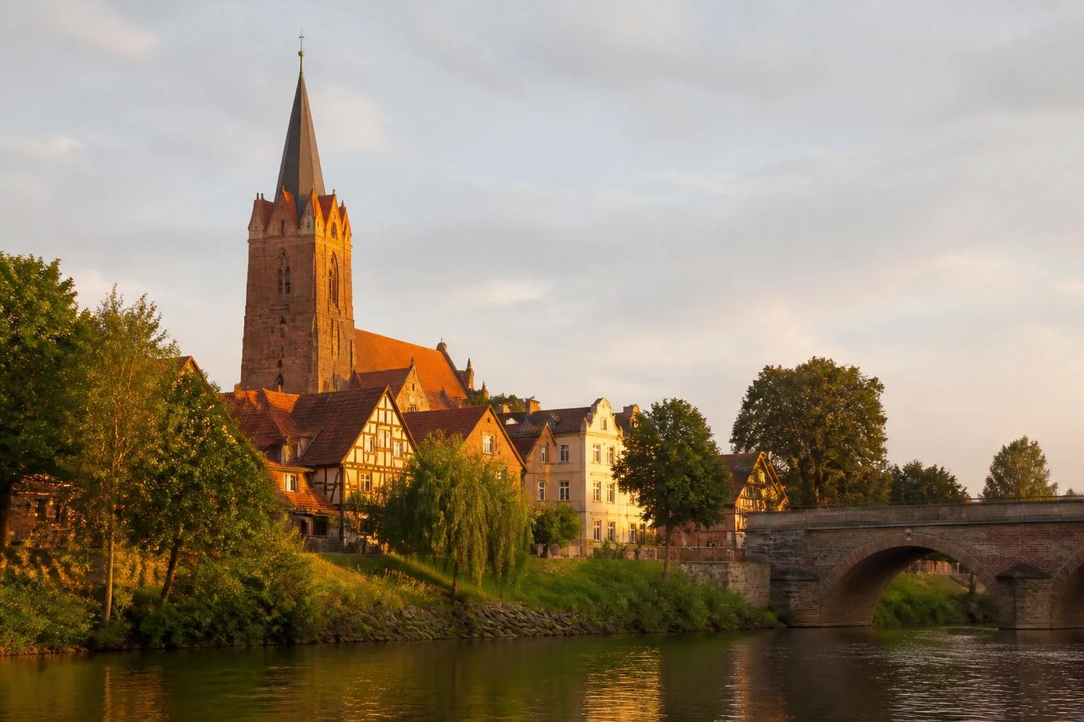 Backsteinkirchturm und Fachwerkhäuser über einem grünen Flussufer mit steinerner Bogenbrücke