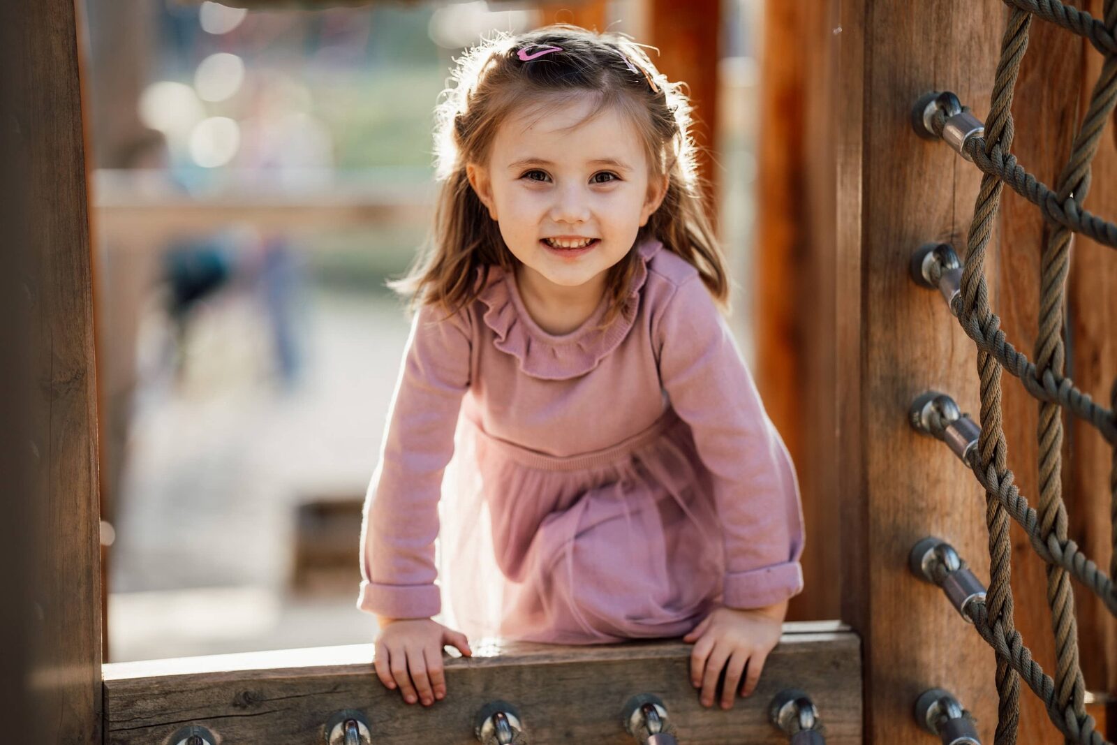 Kindergartenfoto: Lachendes Kita-Kind im rosa Kleid am Klettergerüst, natürliches Porträt im Gegenlicht