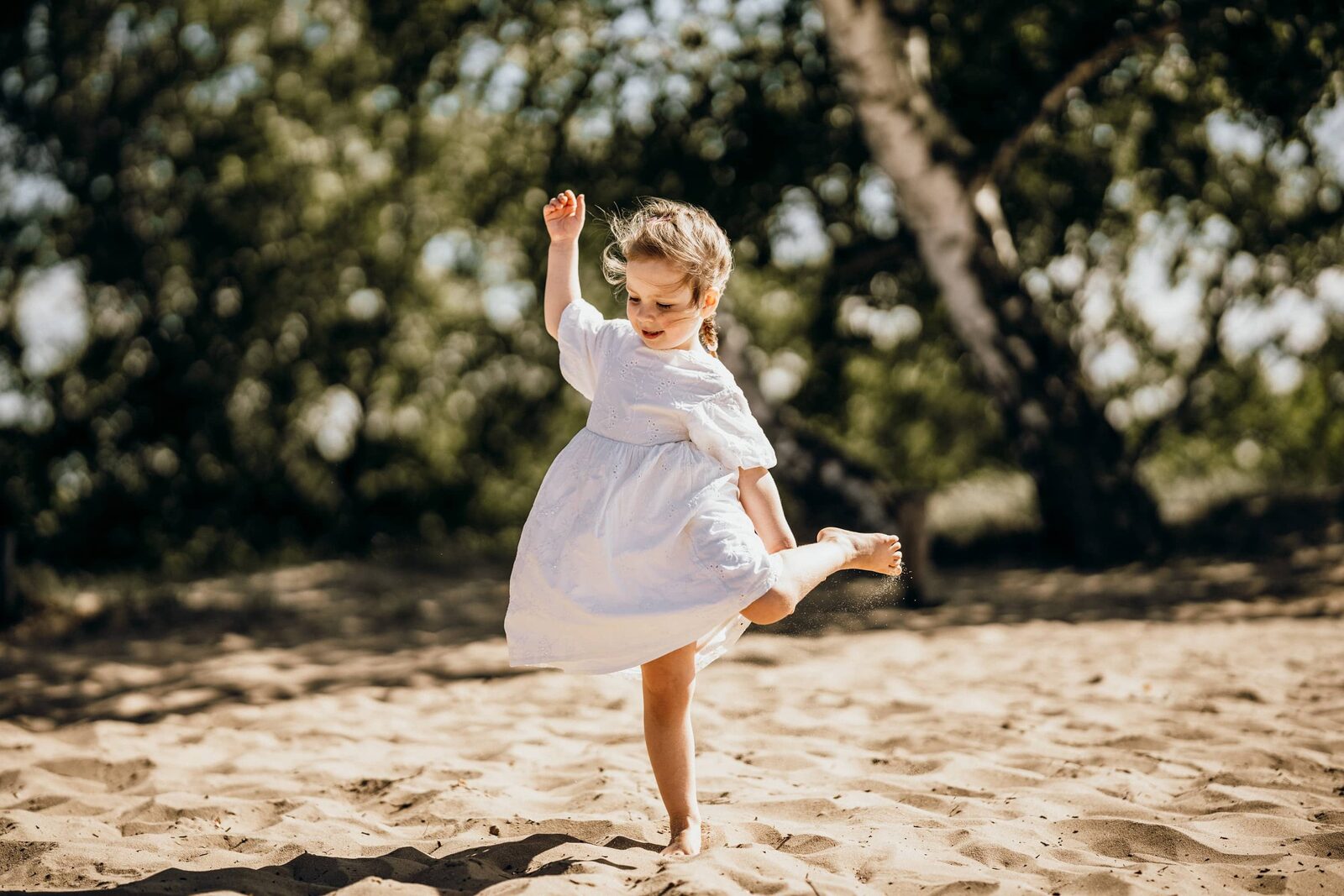Kita-Fotografie: Mädchen im weißen Kleid dreht sich barfuß im Sand unter Bäumen